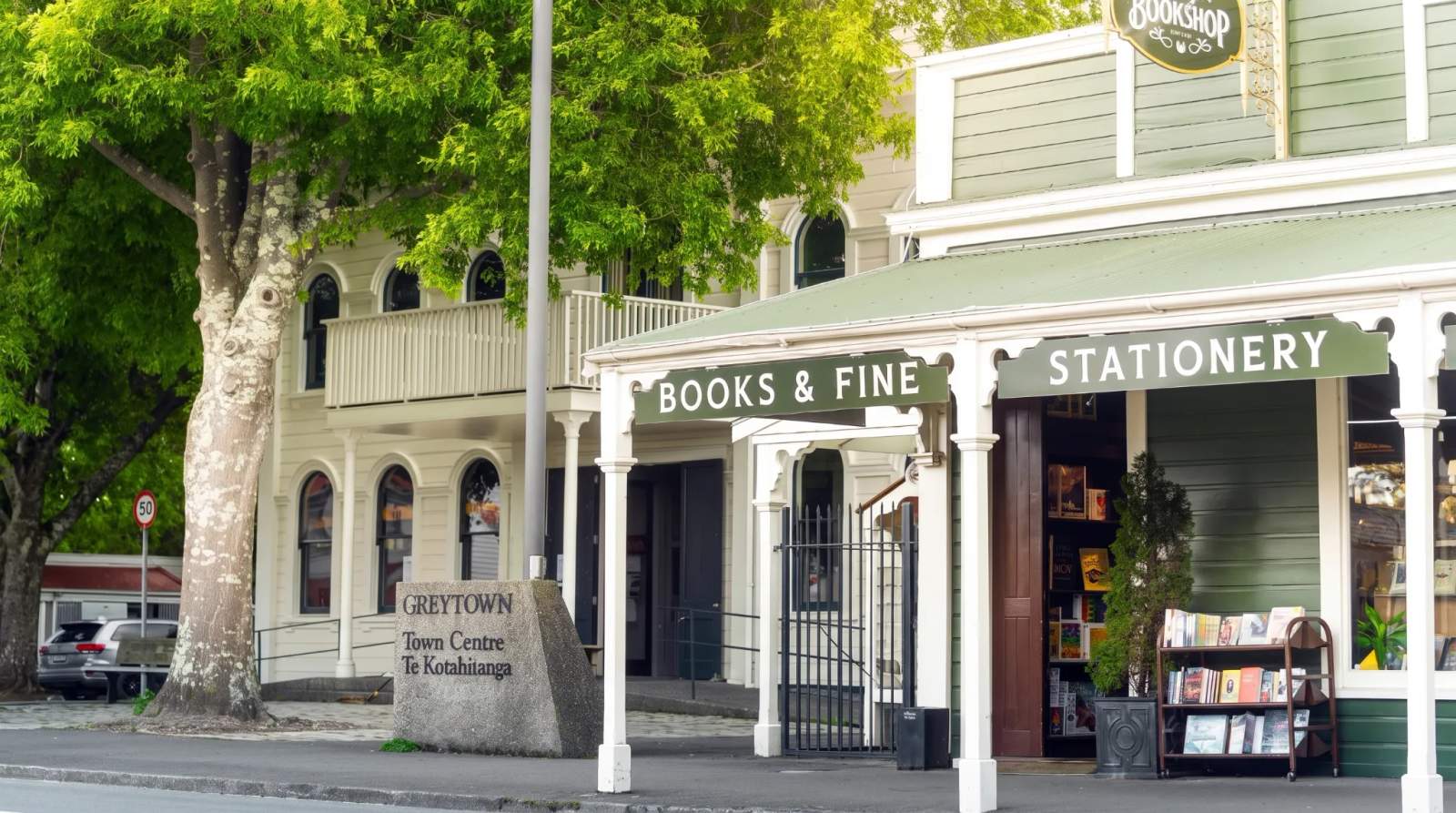 Historic Greytown buildings with green roofs, including a bookstore and a town center sign, under bright green trees.