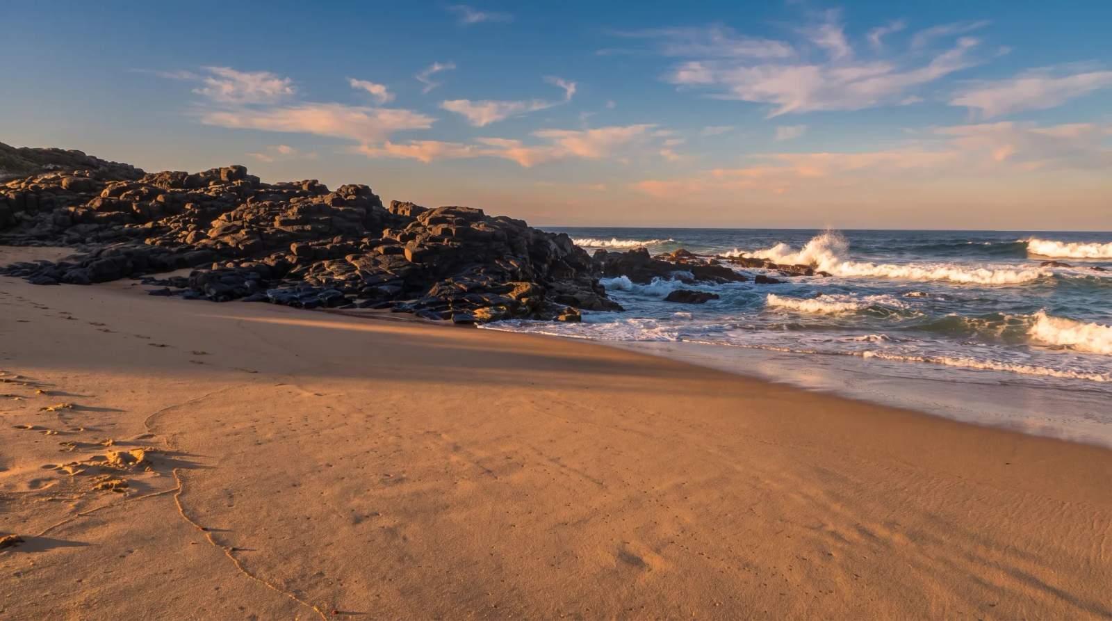 Waves crash on a sandy Blythedale beach with dark rocks under a blue and cloudy sky.