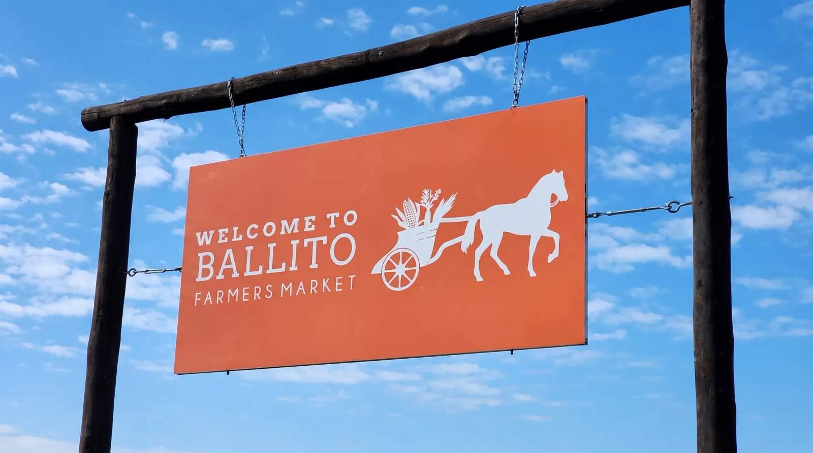 An orange sign welcomes visitors to the Ballito Farmers Market, featuring a horse pulling produce under a blue sky.
