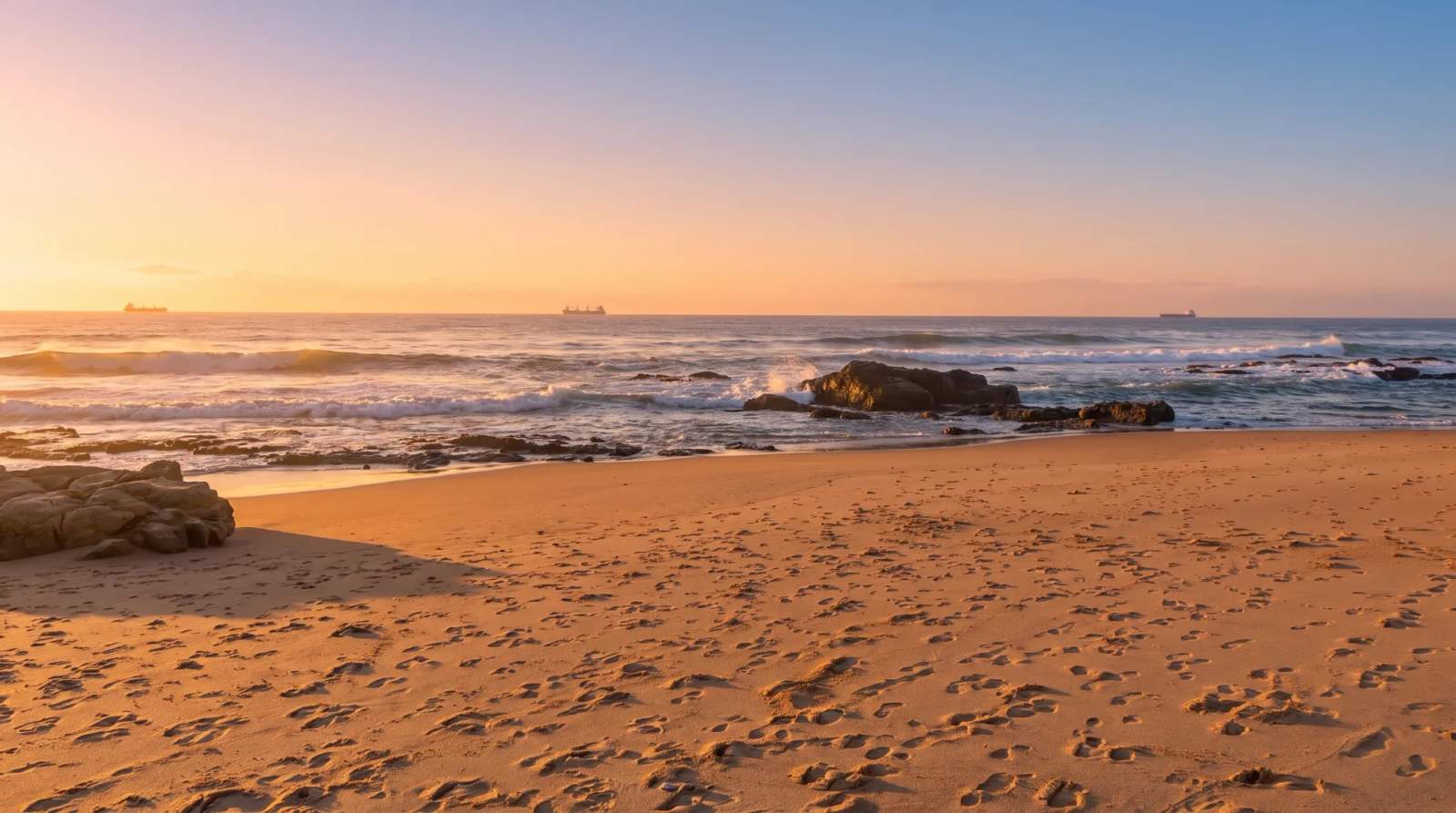 Sunrise over the sandy, rocky beach with ocean waves in Blythedale, with ships on the horizon.