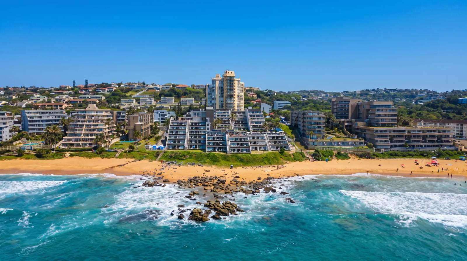 Aerial view of Ballito's coastline, with buildings rising behind a sandy beach, rocks, and blue ocean waves.
