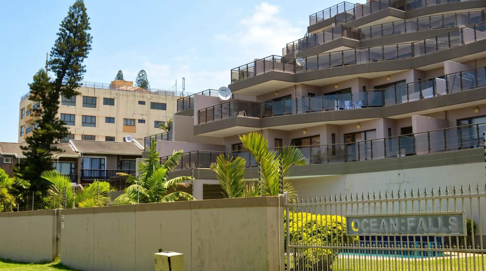 Modern terraced apartments with glass balconies and palm trees in Blythedale, with a fence and wall in front.